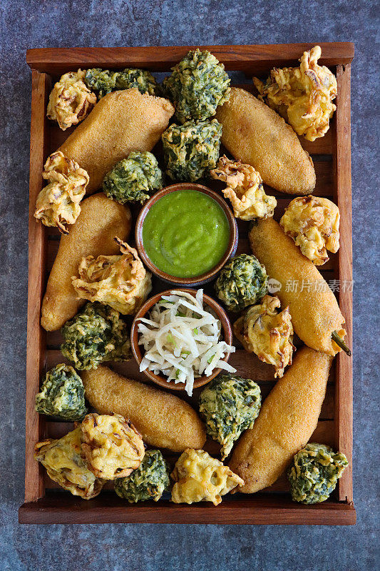 特写画面:木盘自制青椒油饼(mirch pakora)，酥脆的洋葱bhajis，菠菜油饼(palak pakora)，几碗薄荷和香菜酸辣酱，磨碎的奶酪，印度美食素食小吃，灰色背景，高架视野